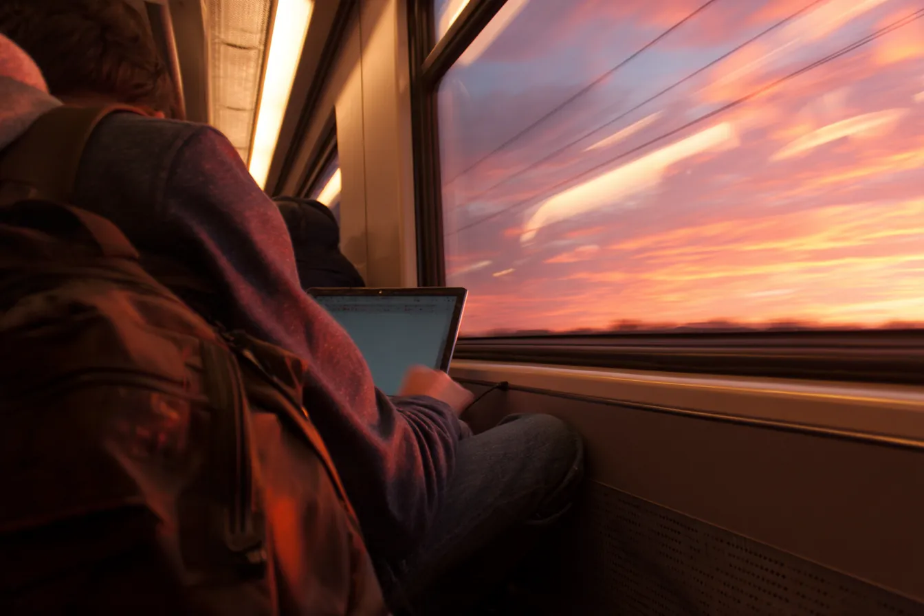 On a commuter train at sunset, a passenger works on a laptop by the window, backpack on the seat beside him.