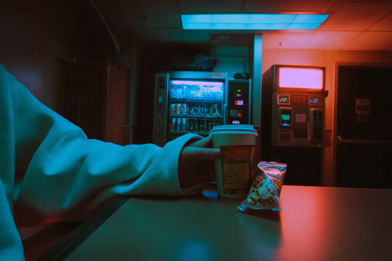 A hospital break room late at night: an arm resting on the table, a paper coffee cup and a snack, vending machines glowing behind.
