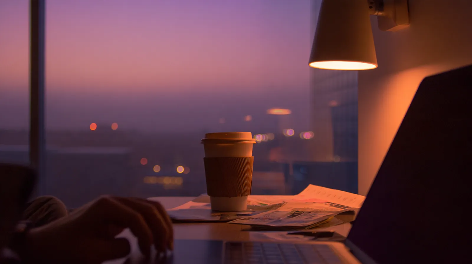A desk by a tall window at dusk: a hand at a laptop, a paper coffee cup and a folded printout under a lamp, city lights blurred beyond the glass.