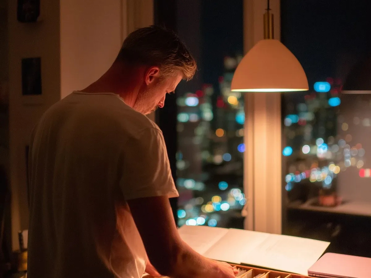 A man in a T-shirt at night working through a drawer of files under a pendant lamp, city lights through the window beside him.